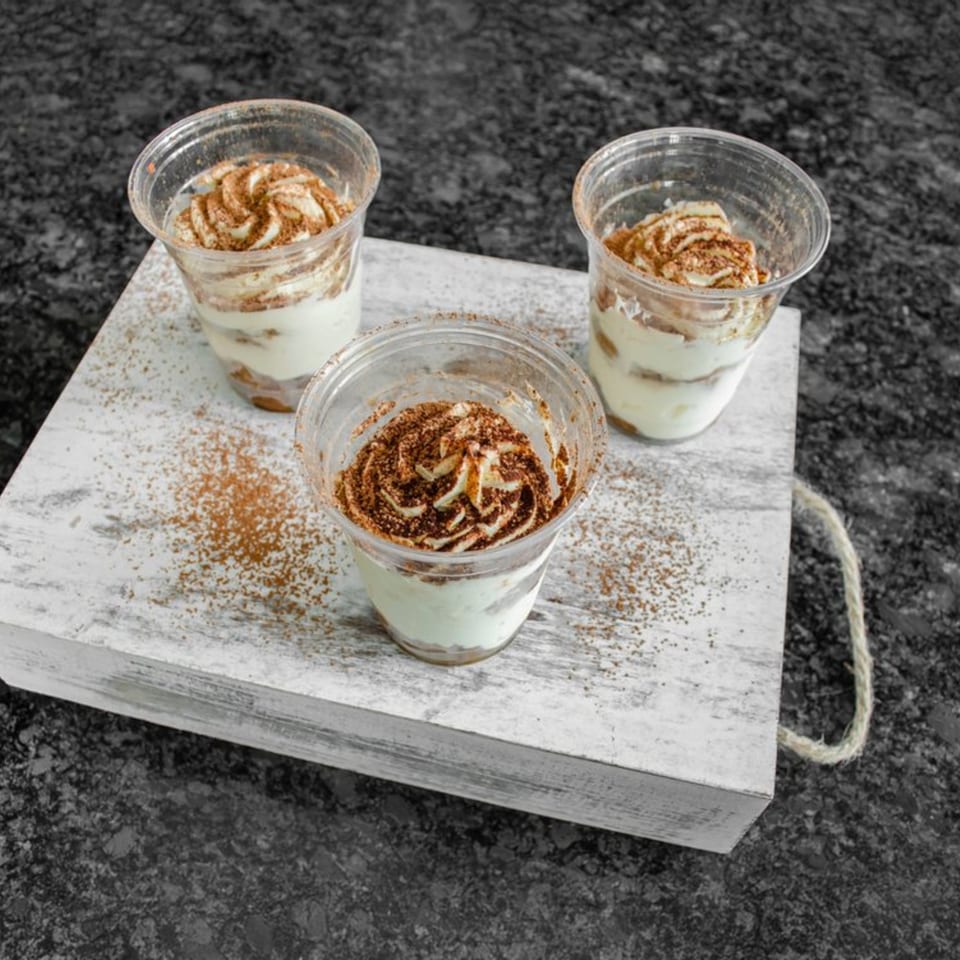Three Bella Ciao tiramisu desserts in clear cups with cocoa powder on a white wooden tray.
