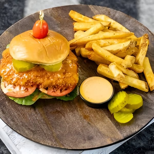 Crispy chicken sandwich with lettuce, tomato, pickles, fries, and dipping sauce on a wooden board.